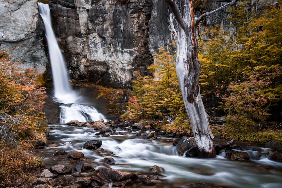 Una cascada que aboca a un riu envoltat de fullatge d'octubre i penya-segats rocosos.