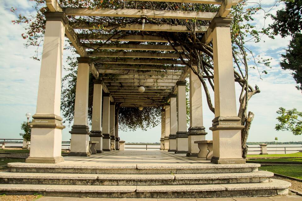 A stone pergola with steps leading up to it, surrounded by greenery and a clear sky.