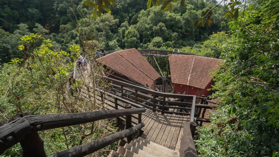 A wooden bridge with a tiled roof leading to a red building surrounded by dense forest.