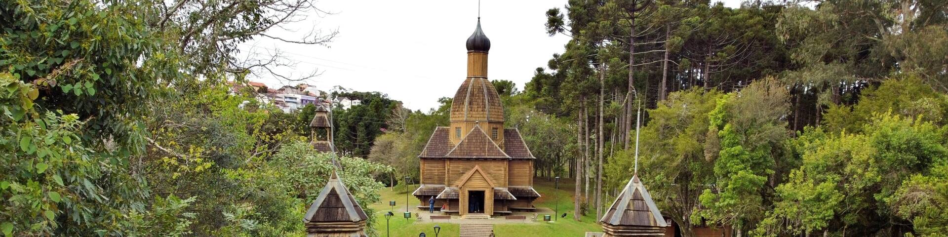 A wooden structure with a pointed roof and two towers, surrounded by lush greenery and a paved pathway leading to it.