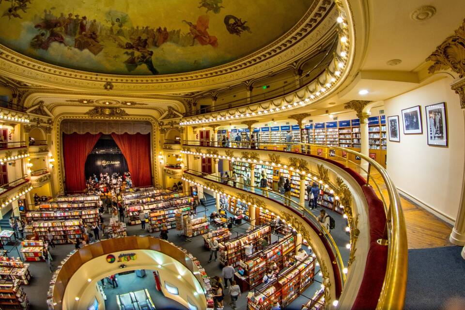 A grand, ornate library with multiple levels of bookshelves and a central stage.