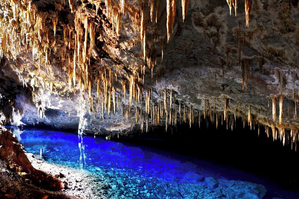 Una cueva con estalactitas colgando del techo y una piscina de agua azul cristalina en el fondo.