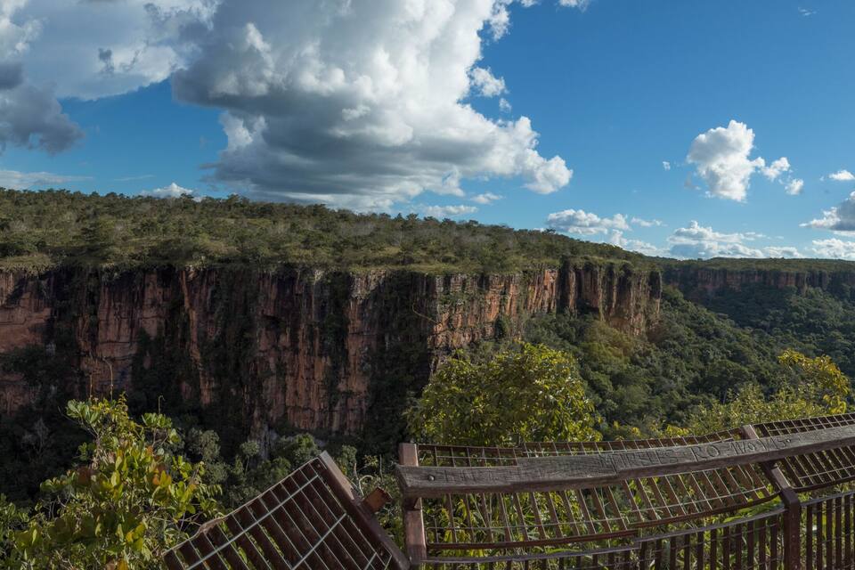 Una cascada se precipita por un acantilado, una plataforma de madera para observar el paisaje, y una cordillera boscosa bajo un cielo parcialmente nublado.