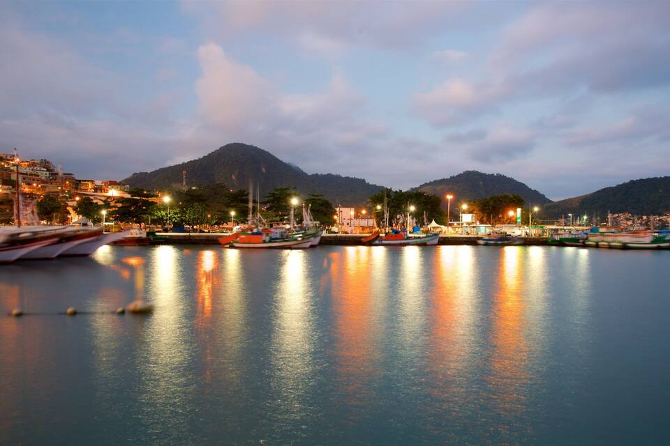 Un puerto al atardecer con barcos amarrados, una montaña en el fondo y luces de ciudad reflejándose en el agua.