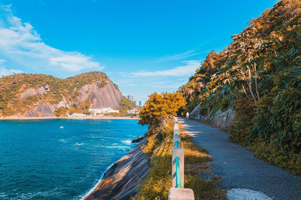 A coastal road with a view of the ocean, a mountain, and a building in the distance.