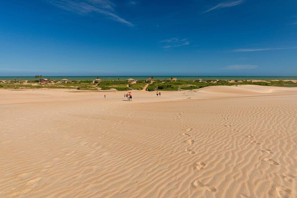 Paesaggio desertico con dune di sabbia, cielo azzurro limpido e edifici in lontananza.