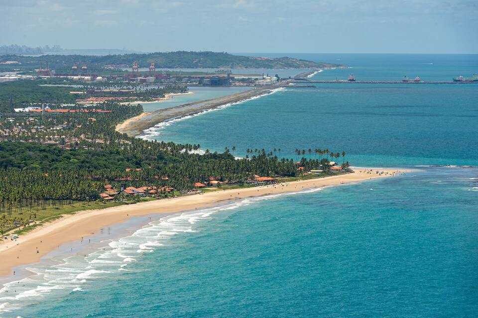 Paisagem costeira com praia, oceano, estruturas industriais e uma linha de costa distante.