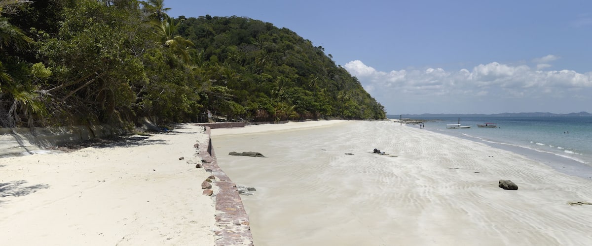 A sandy beach with a rocky shoreline, a forested hill, and calm sea with boats.