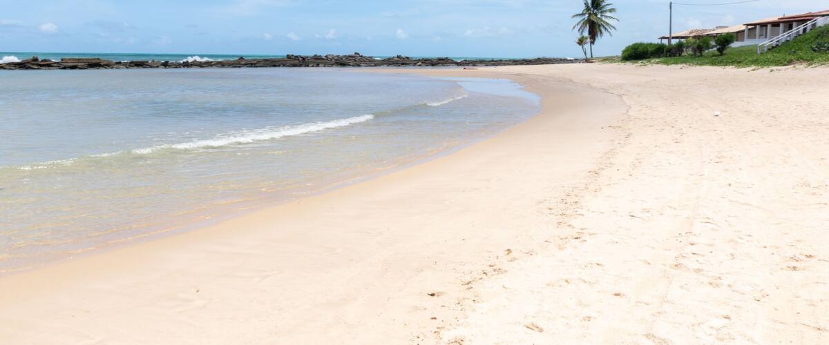 A sandy beach with gentle waves, a palm tree, and a building with a red roof.
