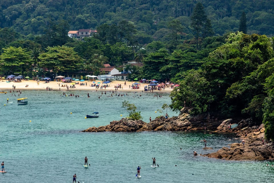 Een druk strand vol mensen die zwemmen en zonnebaden, omgeven door weelderige begroeiing en rotsformaties.