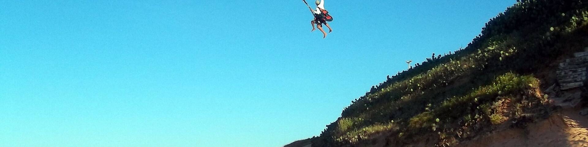 A person paragliding over a sandy beach with tire tracks and a grassy hill in the background.