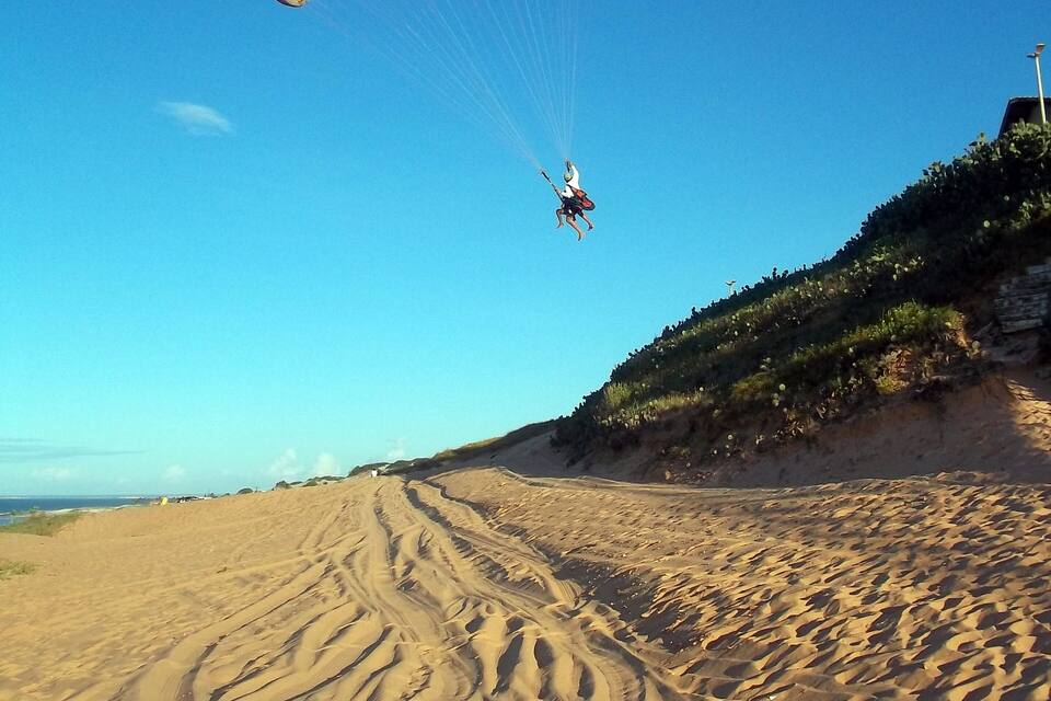 Een persoon paraglidet boven een zandstrand met spoorwielen en een heuvel begroeid met gras op de achtergrond.