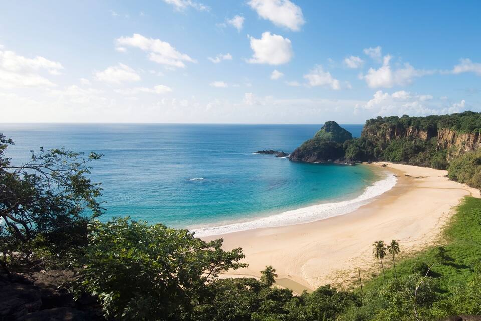 A coastal landscape with a sandy beach, a clear blue sea, and a forested cliff.
