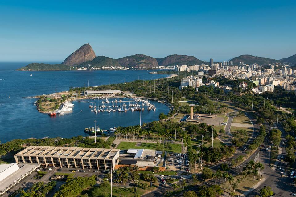 Paisagem costeira com um porto, uma montanha imponente e edifícios urbanos densos.