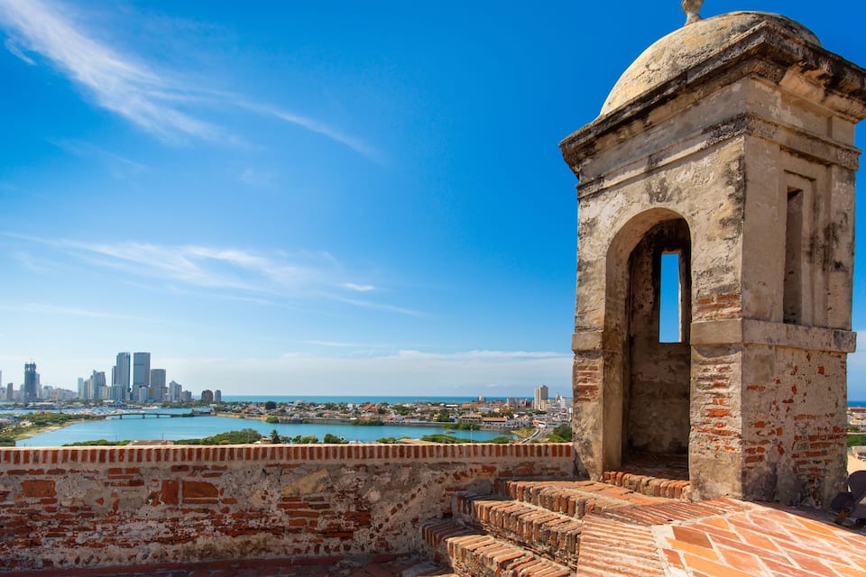 A historic stone tower with a dome, overlooking a coastal cityscape with a bridge and a clear blue sky.