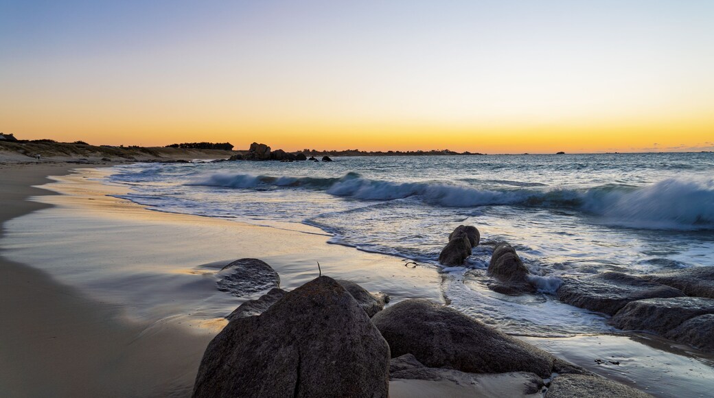 La plage des Amiets s’embrase sous un ciel orangé et doré. Les vagues laissent des reflets scintillants sur le sable mouillé, tandis que les rochers se découpent dans cette lumière crépusculaire.