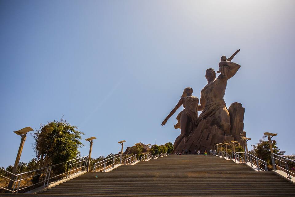A large outdoor sculpture with multiple figures, a staircase leading up to it, and a clear blue sky.