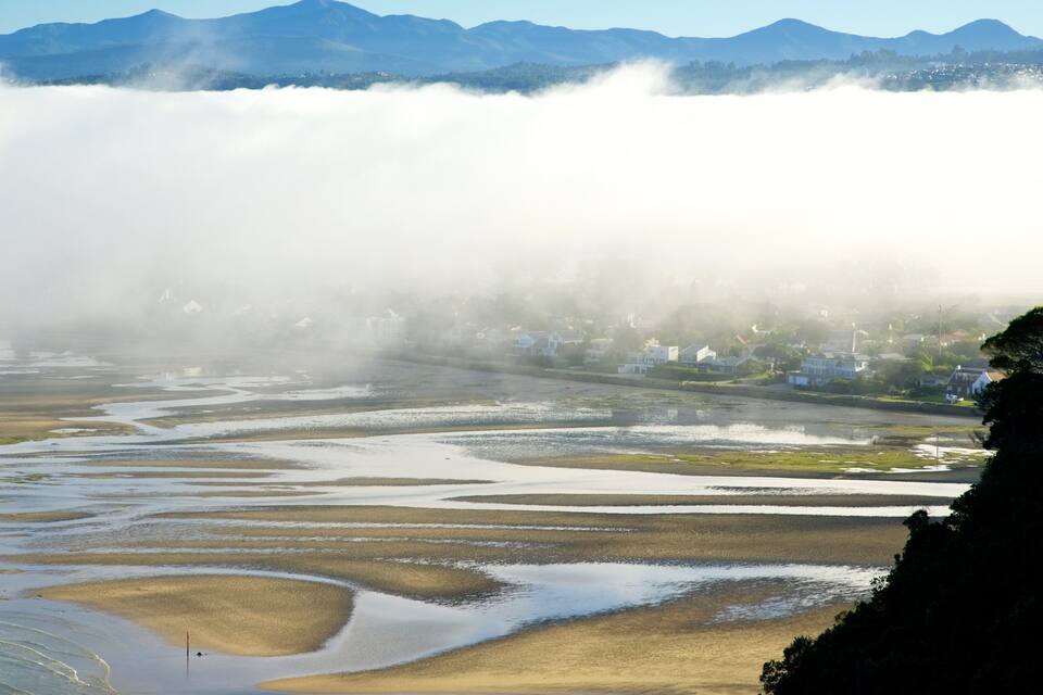 A geothermal area with steam rising from the ground, surrounded by a river with wet sand and a town in the background.