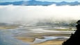 A geothermal area with steam rising from the ground, surrounded by a river with wet sand and a town in the background.