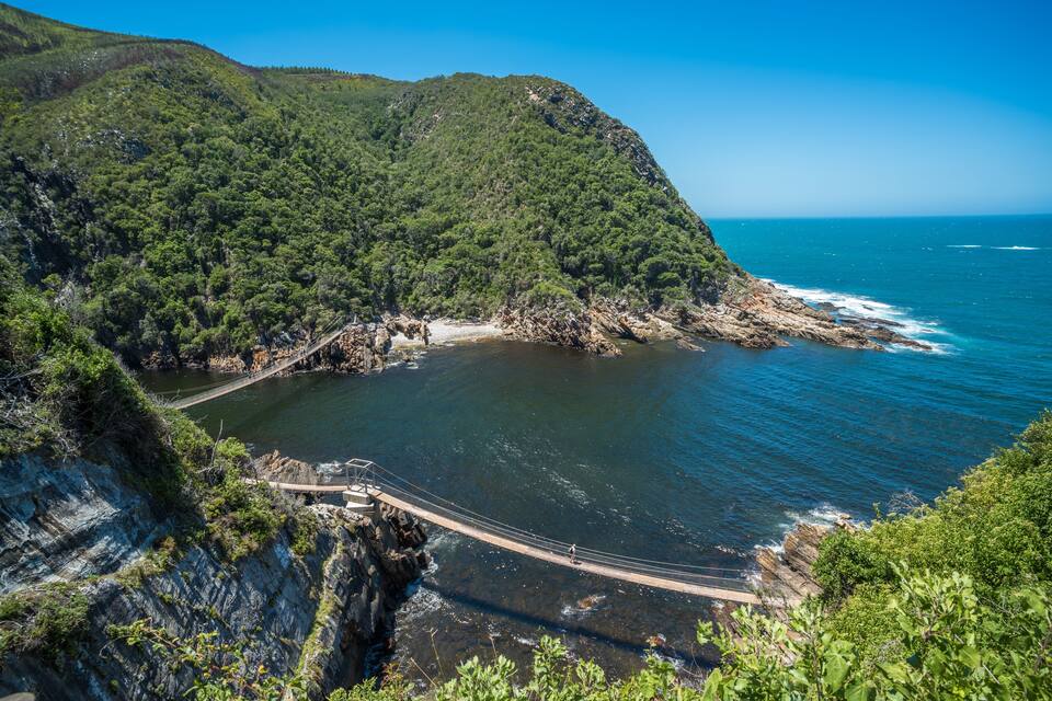 A coastal landscape with a suspension bridge, rocky cliffs, and a clear blue sea.