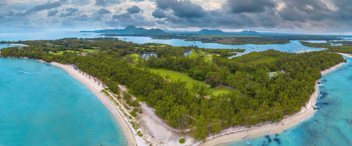 A coastal landscape with a beach, dense forest, and a distant mountain range under a cloudy sky.