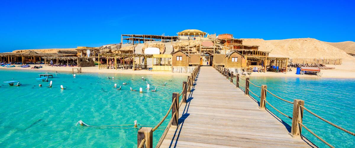 A wooden pier leading to a beach with clear turquoise water, thatched-roof structures, and a sandy shoreline.