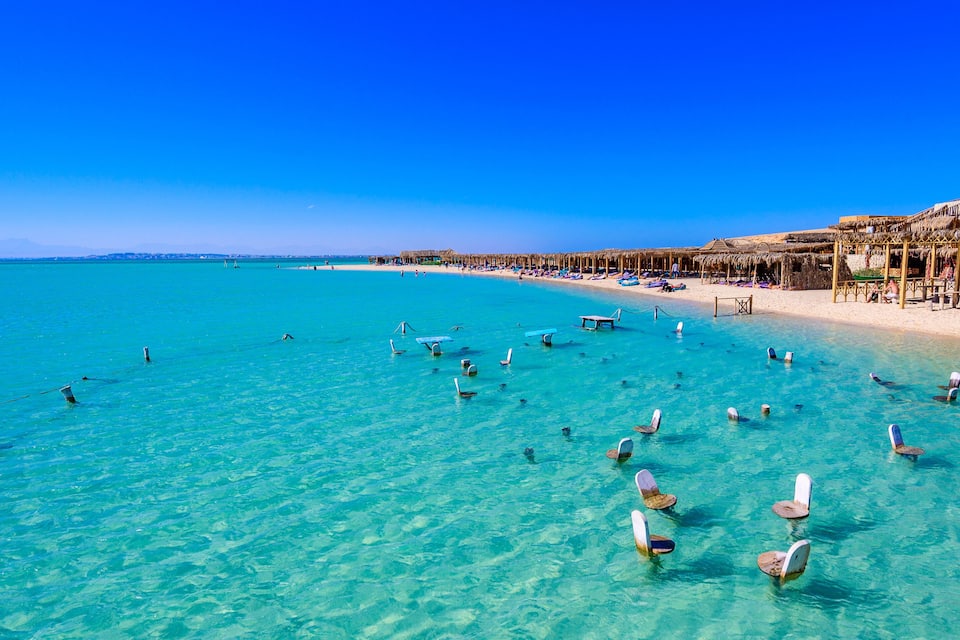 A beach with clear turquoise water, sun loungers, and thatched-roof structures.