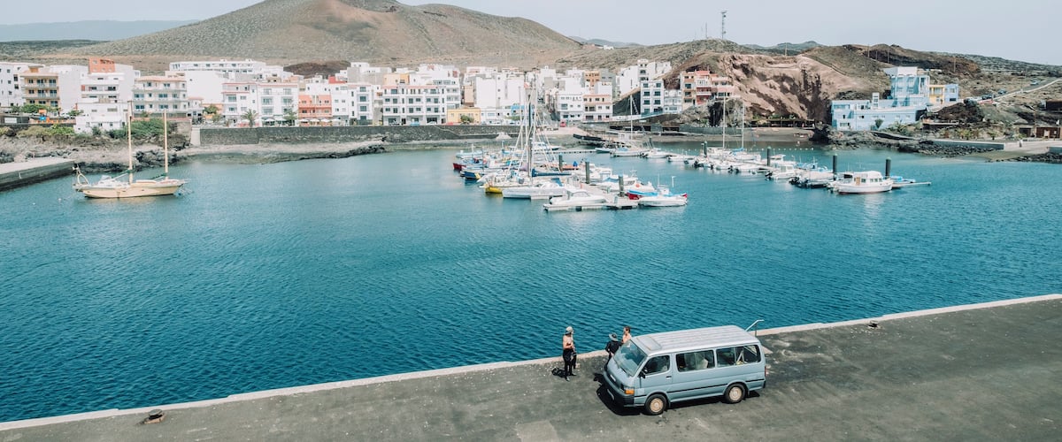 A coastal town with a marina filled with boats, a van parked by the waterfront, and colorful buildings in the background.