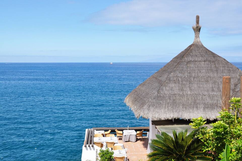 Estructura con techo de paja que da vista al océano, con una mesa y sillas en una terraza.