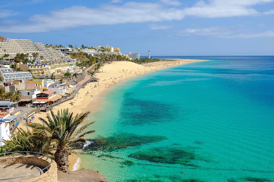 A coastal town with a sandy beach, clear turquoise waters, and a lighthouse in the distance.
