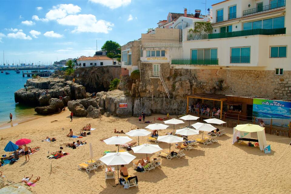 A beach with people sunbathing and relaxing under umbrellas, a rocky coastline, and a bar named Praia da Rainha.