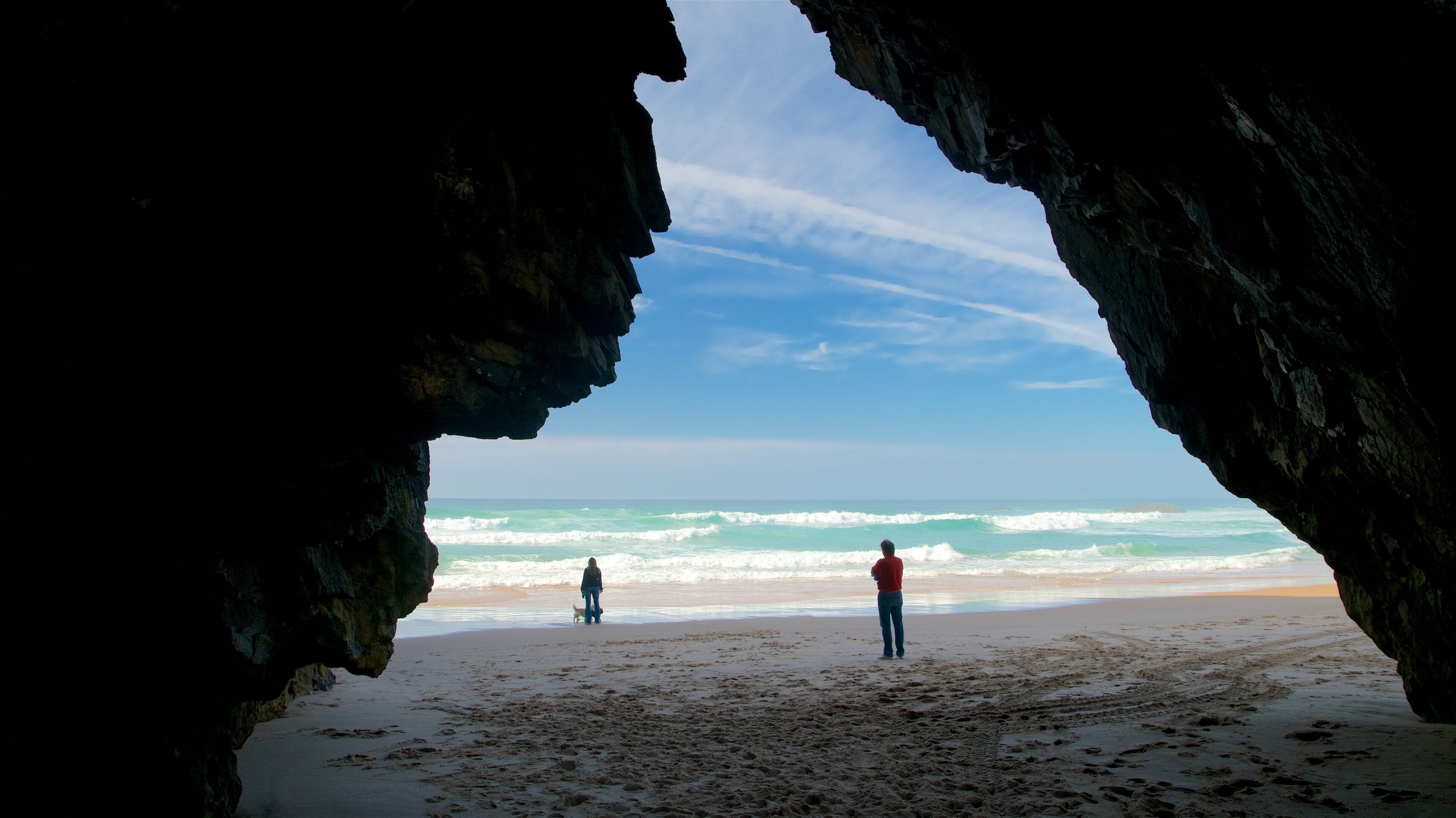 Uma praia com a entrada de uma gruta, ondas e duas pessoas na areia.