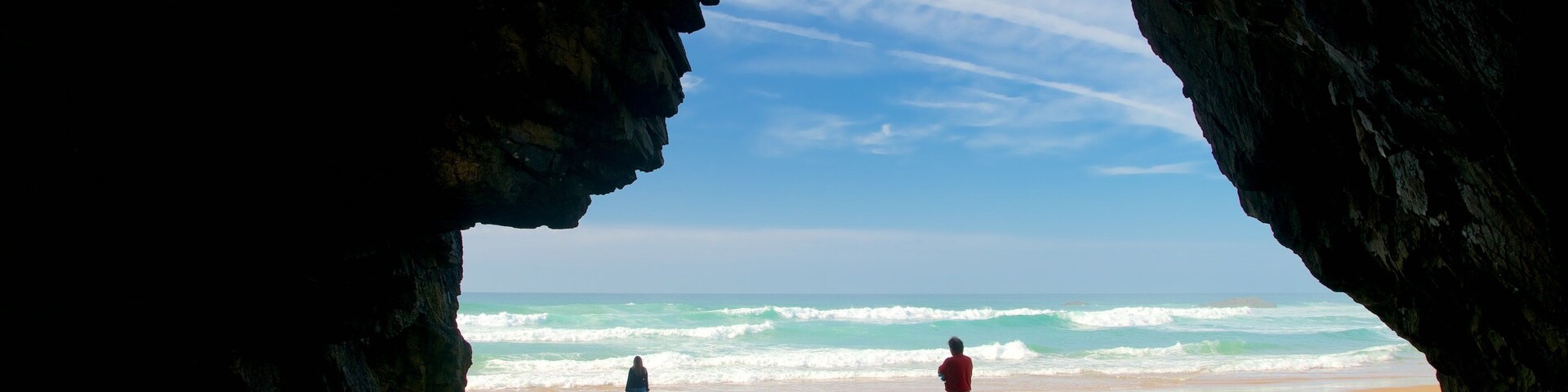 Uma praia com a entrada de uma gruta, ondas e duas pessoas na areia.