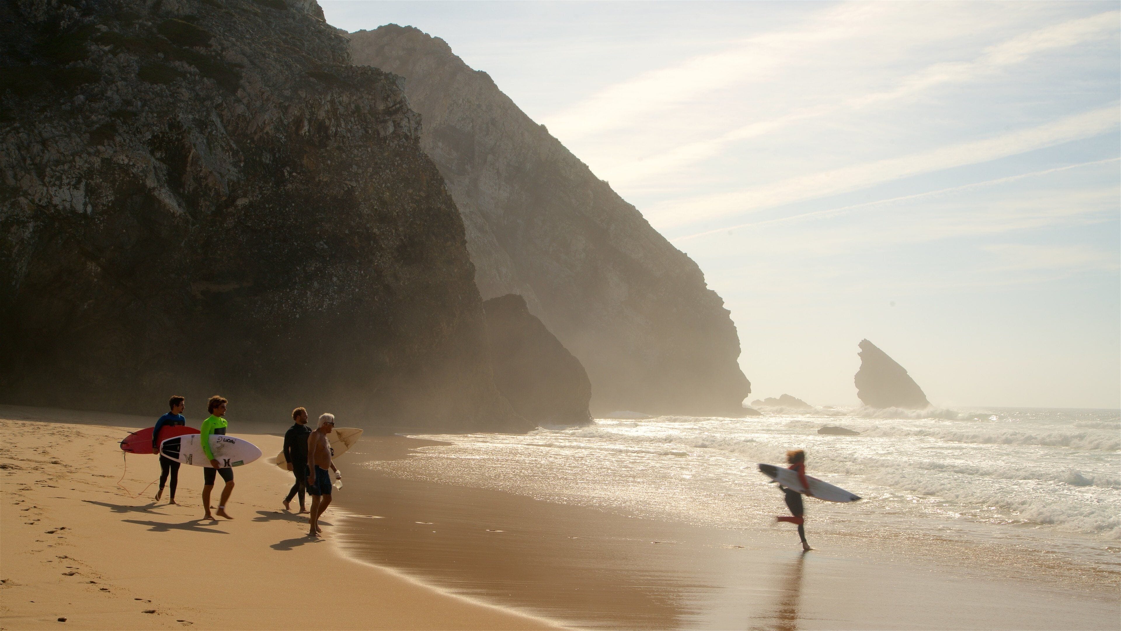 A beach with large rock formations and surfers walking along the shore.