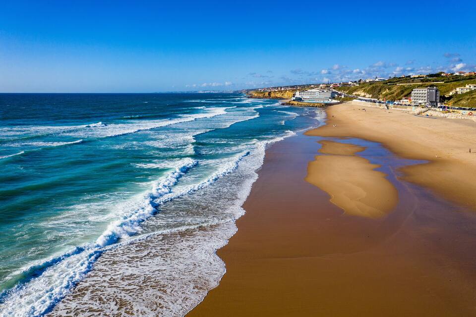 A beach with waves, a sandy shore, and buildings in the background.