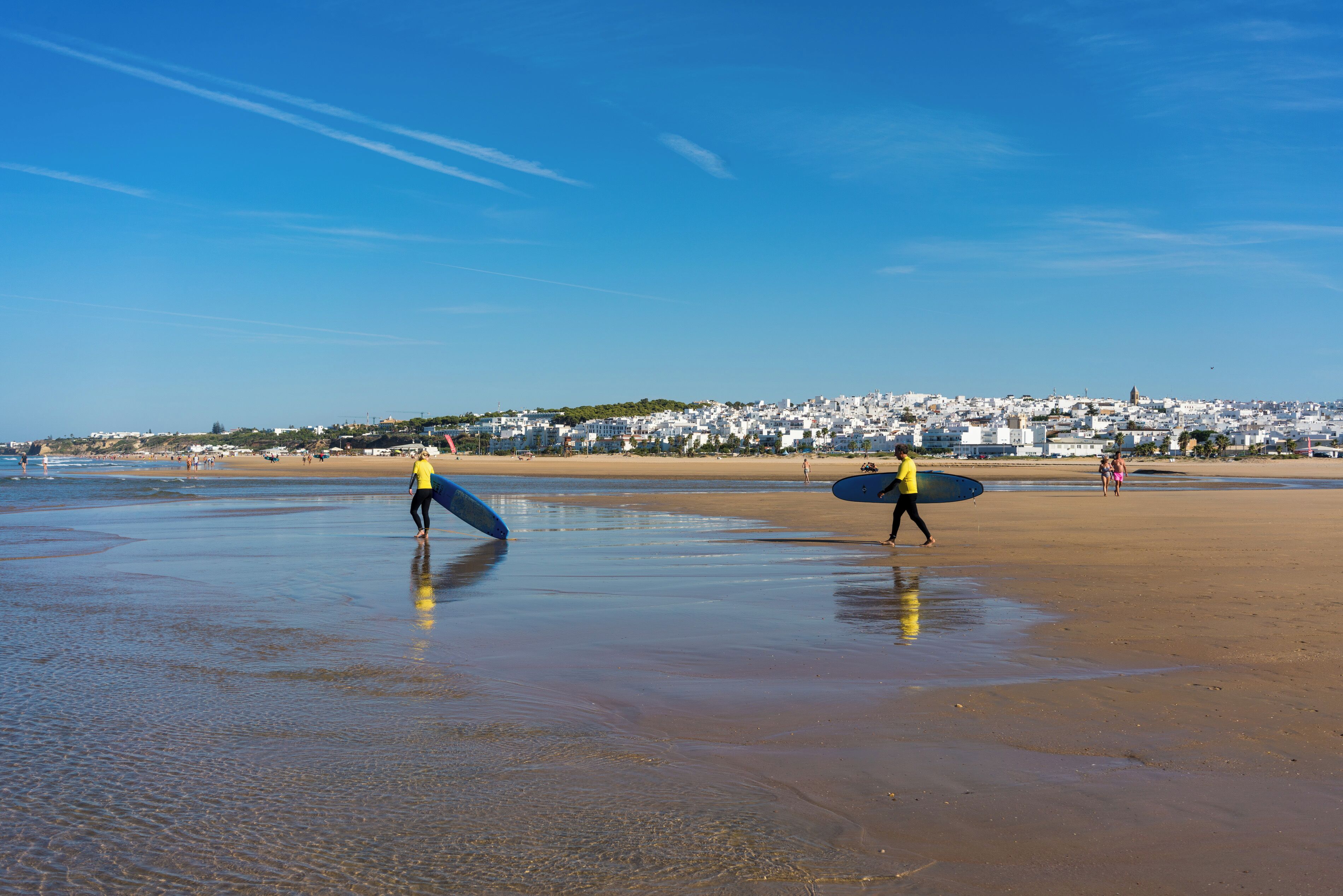 A beach with two people carrying surfboards, a clear blue sky, and a distant cityscape.