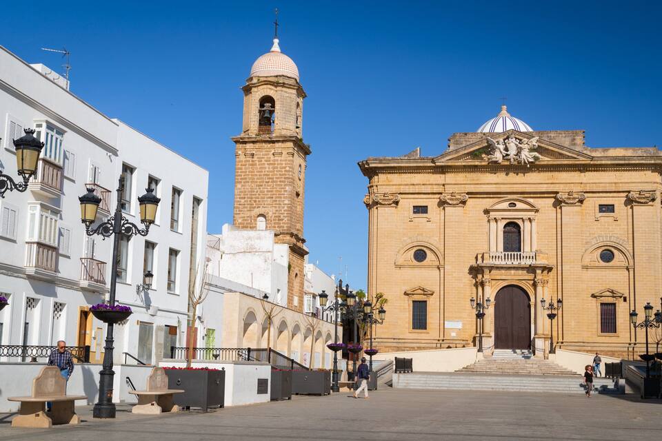 A historic square with a prominent church featuring a bell tower and a dome, surrounded by white buildings with balconies and street lamps.