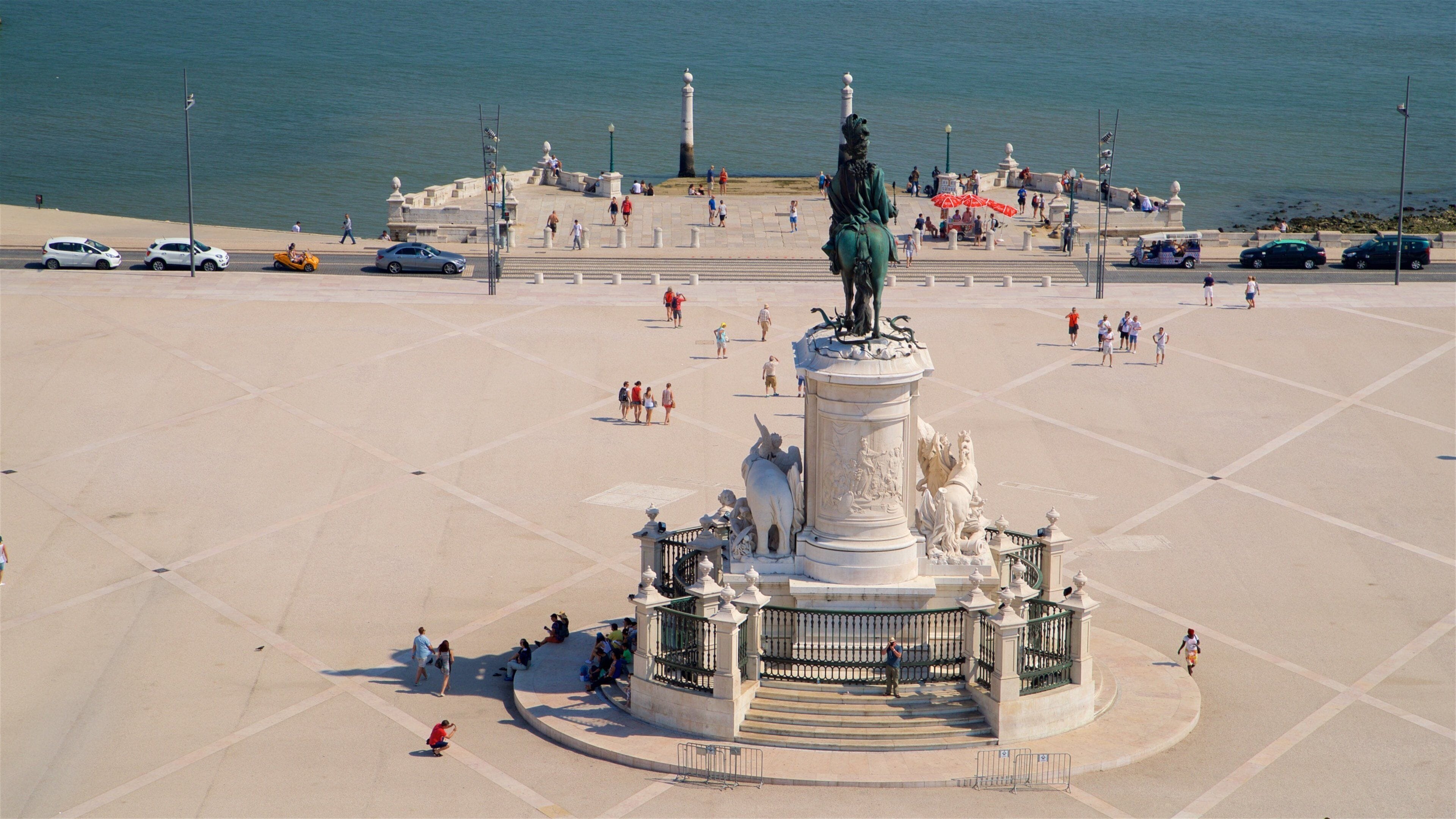 Uma grande praça aberta com uma estátua central, cercada por um muro baixo e escadas que levam a ela.