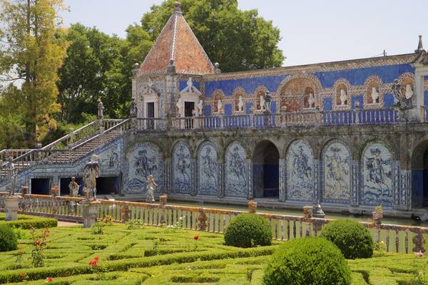 Un bâtiment historique remarquable, orné de motifs complexes en carreaux bleus et blancs, doté d’une tour imposante et agrémenté d’un jardin bien entretenu, avec des haies et des fleurs.