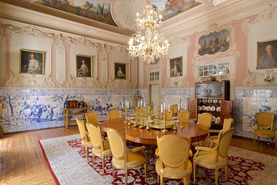 A grand room with ornate decorations, a chandelier, and a large dining table surrounded by chairs.