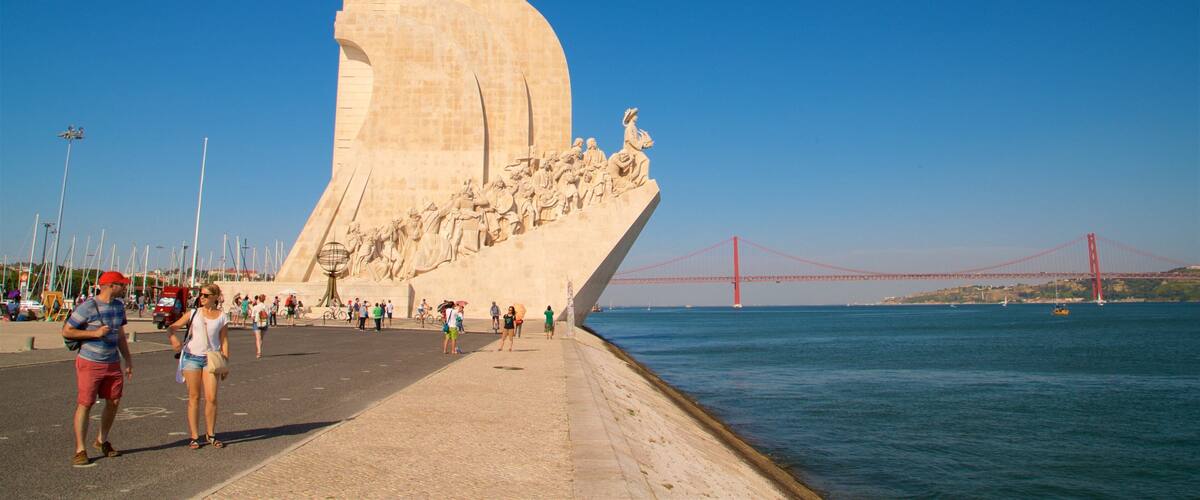 A large monument with a curved base and a group of statues on top, overlooking a body of water with a bridge in the background.