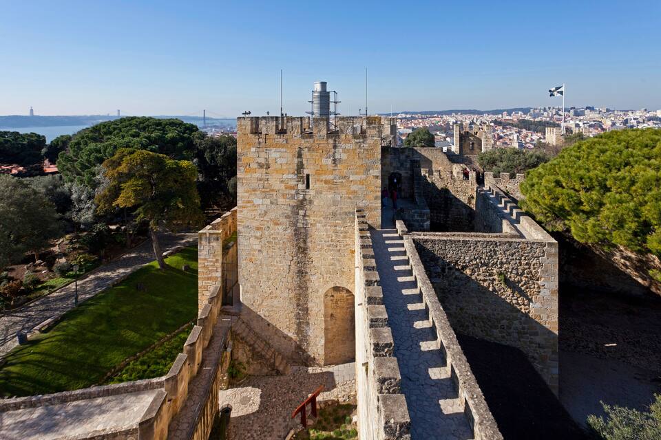 A historic stone tower with a flag, surrounded by greenery and a cityscape in the background.