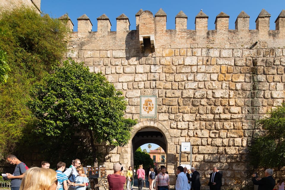 Un muro de piedra con almenas y un escudo de armas, un arco de entrada y gente caminando.