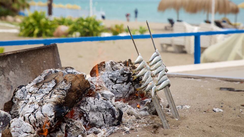 A grill with skewers of fish over charcoal, with a beach and umbrellas in the background.