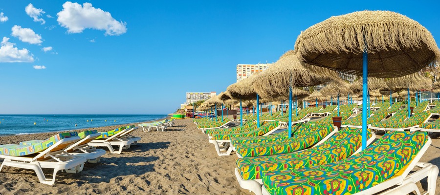 Een strand met rijen kleurrijke ligbedden en rieten parasols.