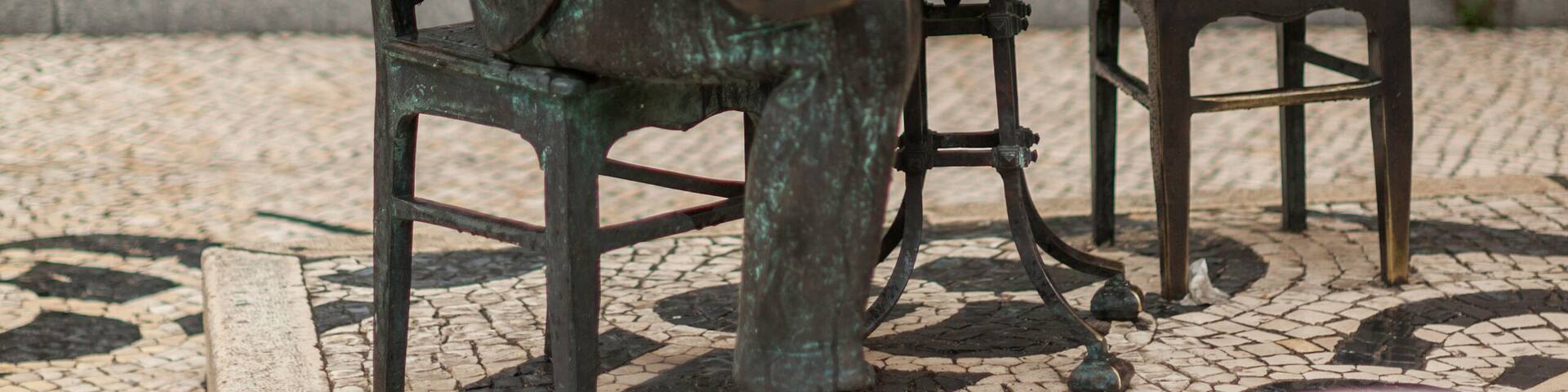 Una estatua de bronce de un hombre con traje y sombrero sentado a una mesa con una silla.