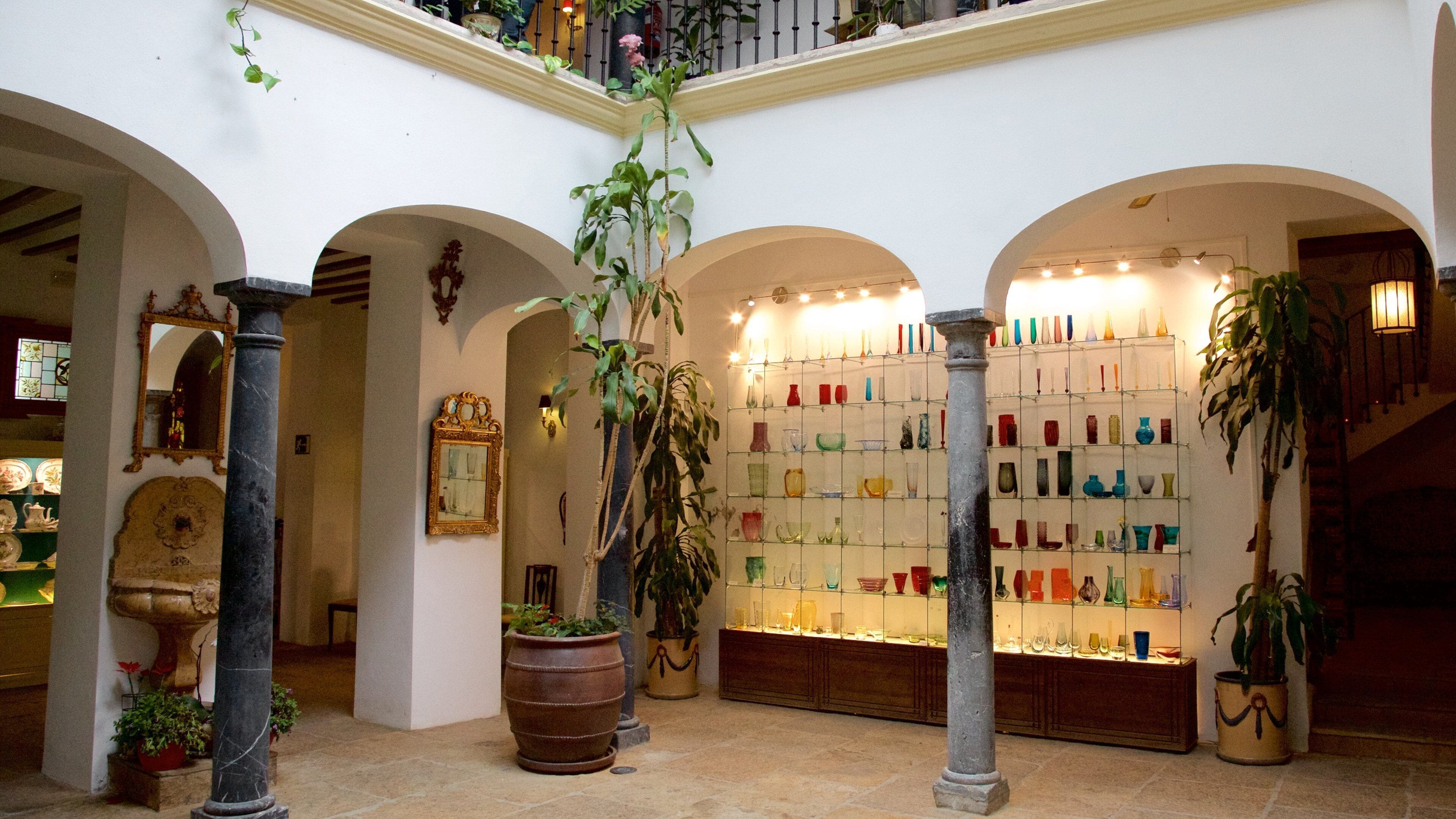 Une galerie où sont exposés des objets en verre, entourée d’arches et de plantes en pot.