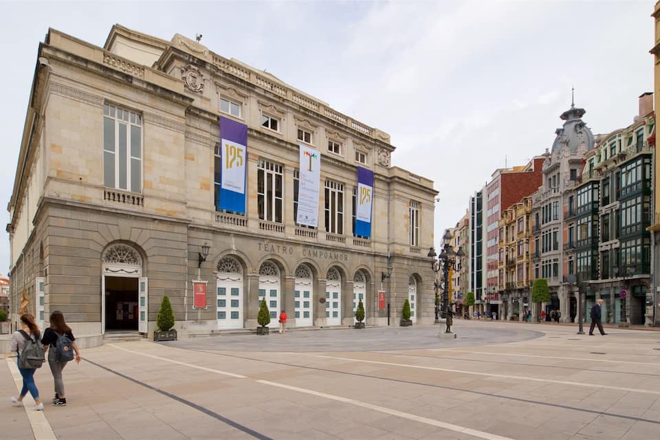 A historic building with arched windows and a central entrance, surrounded by a plaza with people walking.