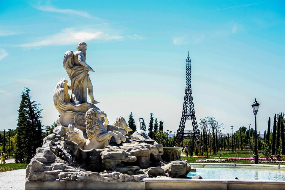 A large outdoor sculpture of a man and woman in a park with a fountain, surrounded by trees and a tall tower.