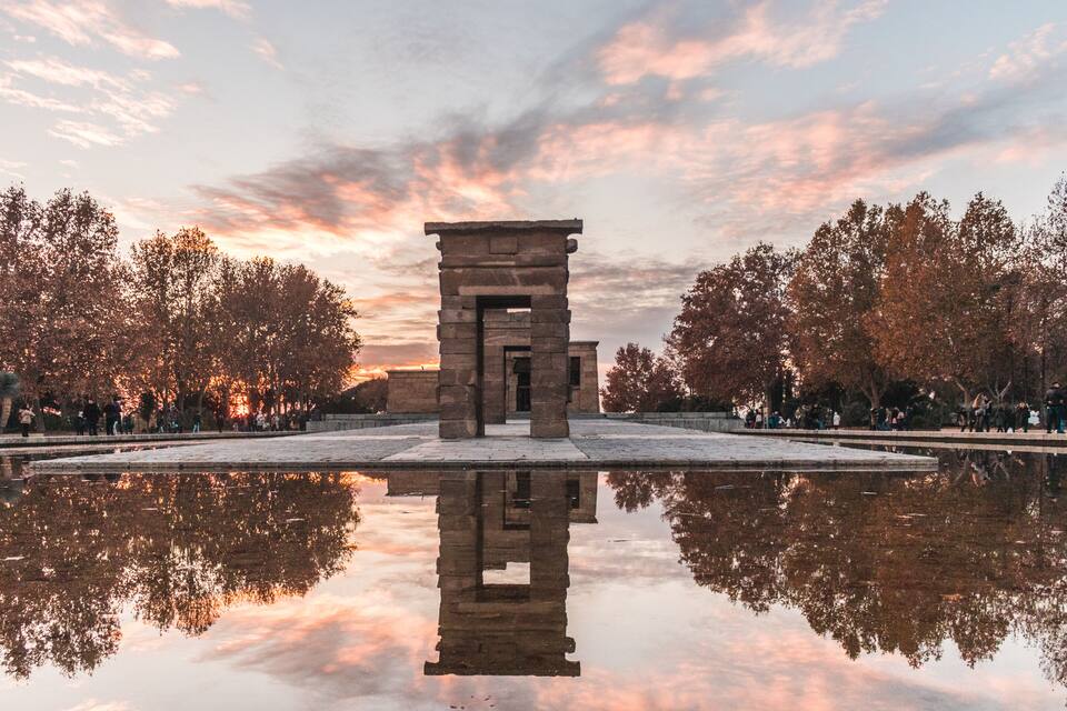 Estructura de piedra con su reflejo en el agua, rodeada de árboles y un cielo al atardecer.